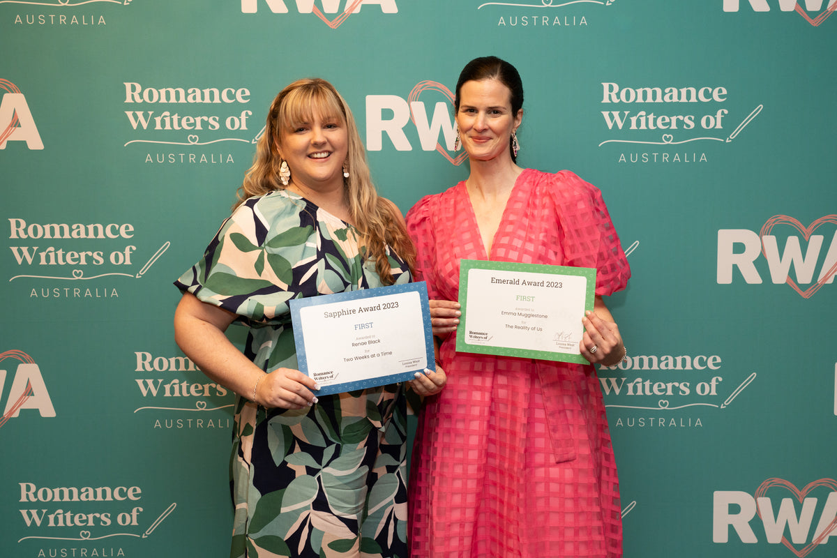 Renae Black and Emma Mugglestone standing in front of a green backdrop featuring the Romance Writers of Australia logo, holding award certificates. One person is wearing a vibrant pink dress with a textured pattern, while the other is dressed in a green and white patterned outfit. Both are smiling as they pose for the photo, celebrating their achievements. The certificates read "Sapphire Award 2023 – First Place" and "Emerald Award 2023 – First Place," recognising their success in writing competitions. 